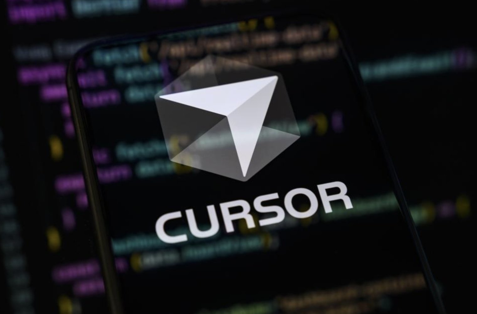 The Cursor logo on a phone screen held up in front of a blurred code editor in the background